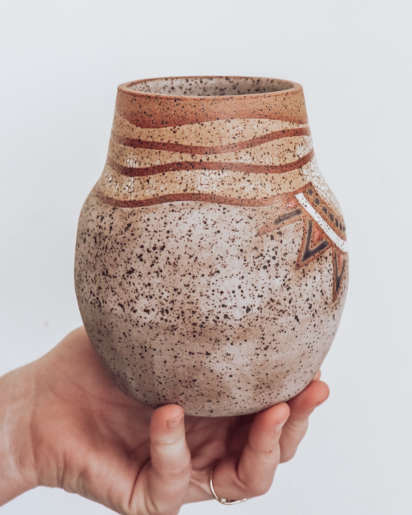 side view of A hand holding a small brown and beige ceramic vase with a crackled texture and geometric patterns.