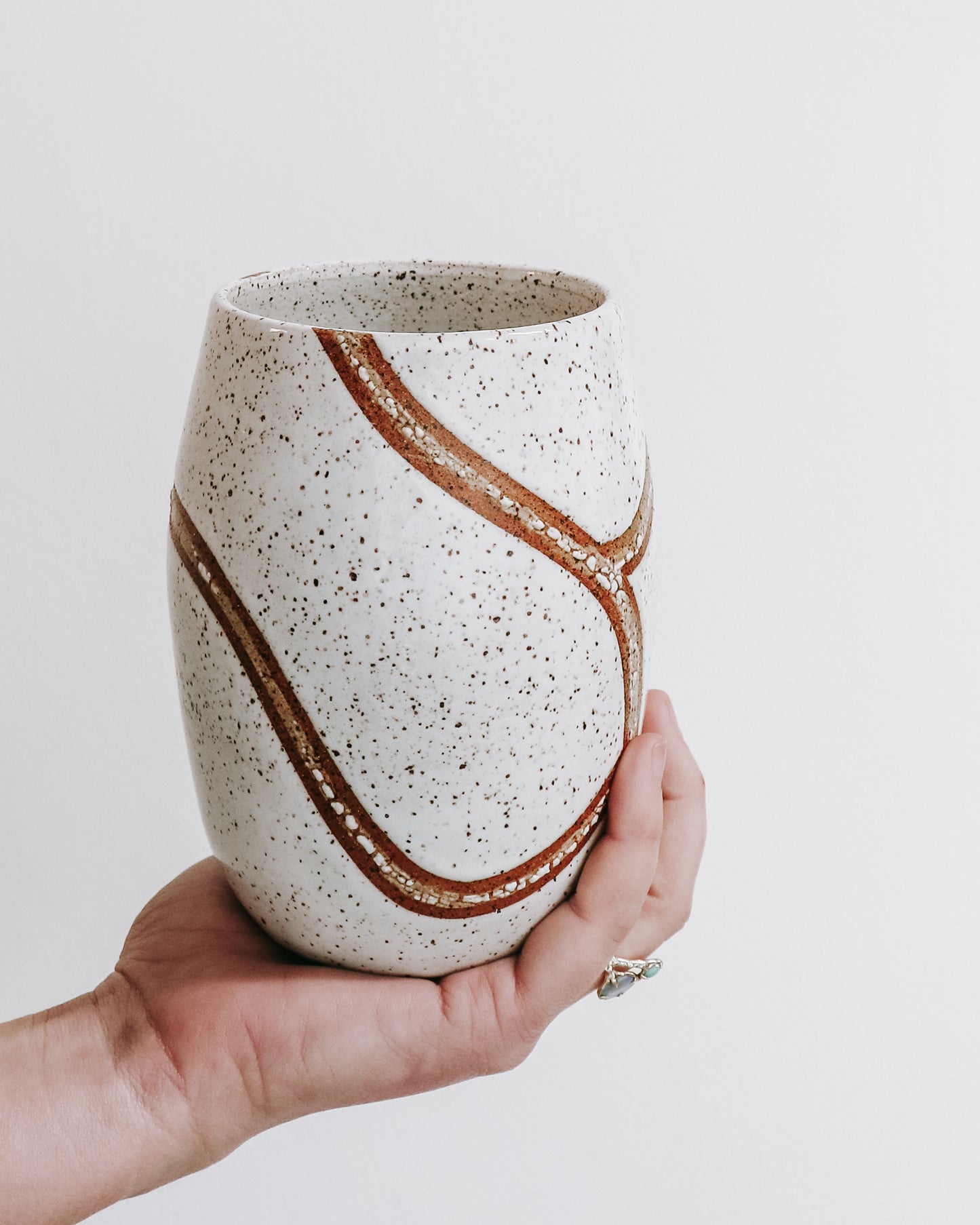 hand holding a handmade speckled ceramic tumbler with flowing carved brown line detailing. 