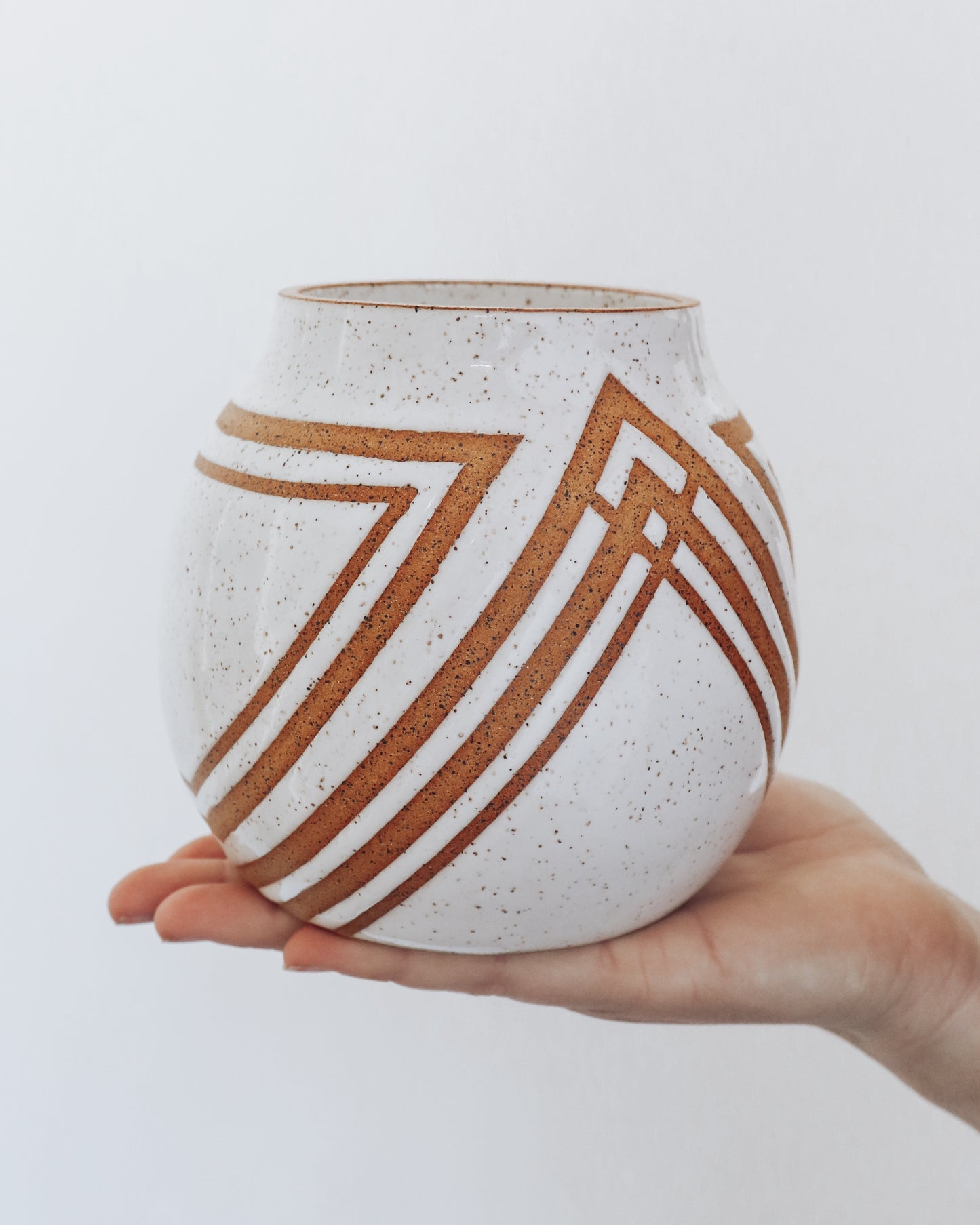  Hand holding a handmade speckled ceramic vase with geometric stripe detailing in warm brown tones. 