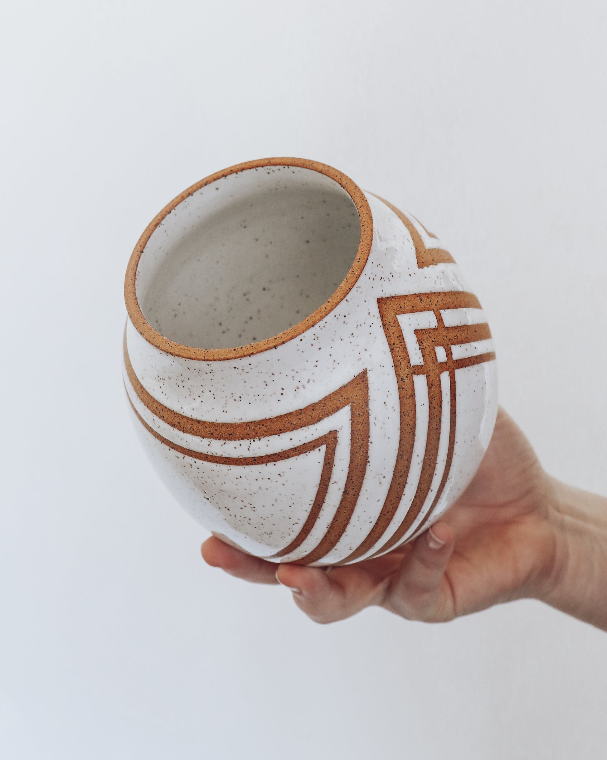  Hand holding a handmade speckled ceramic vase with geometric stripe detailing in warm brown tones. 